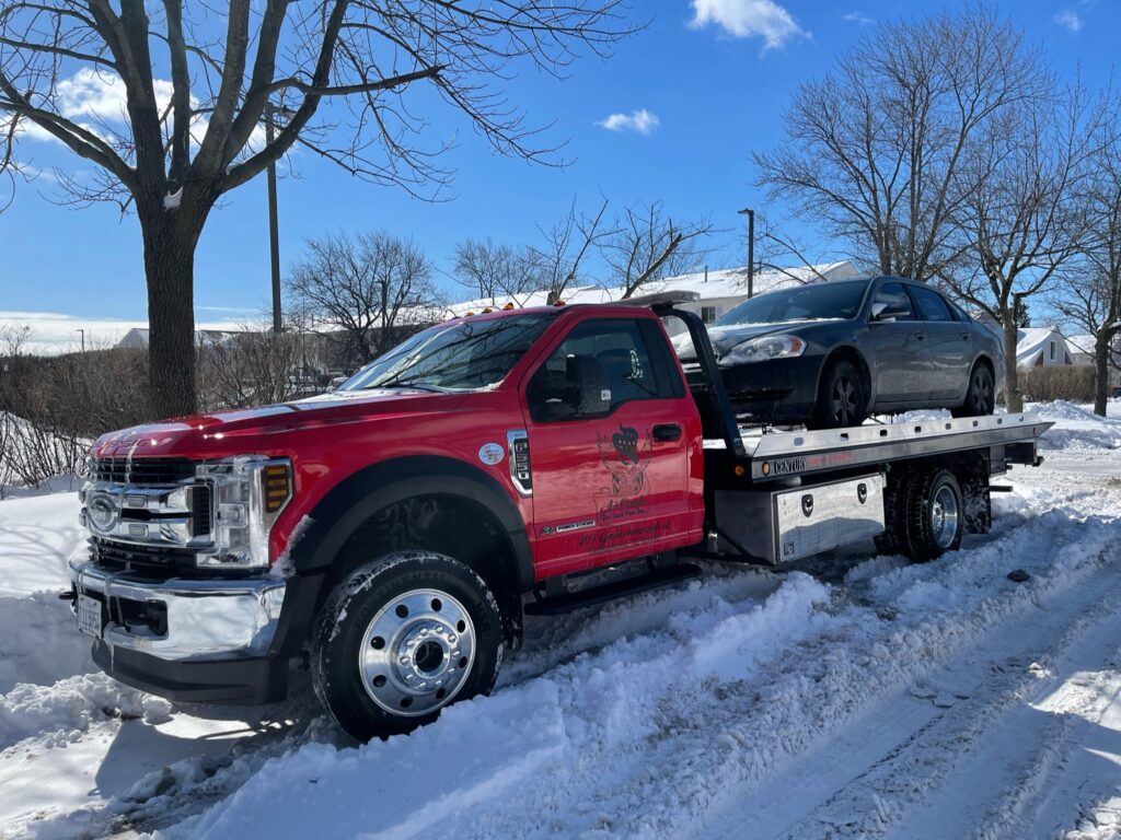 A red tow truck on the snowing street towing away a black junk car