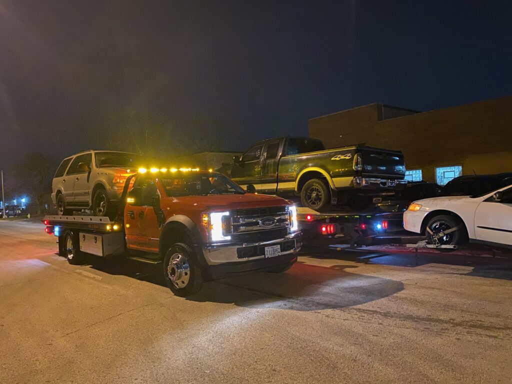 Two tow trucks on the street towing away a gray and black junk car at night