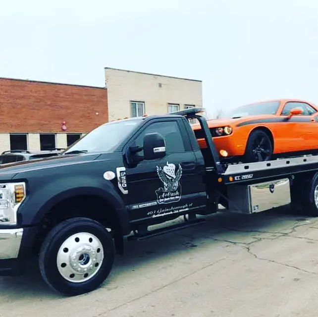A tow truck on the street towing away a orange junk car.