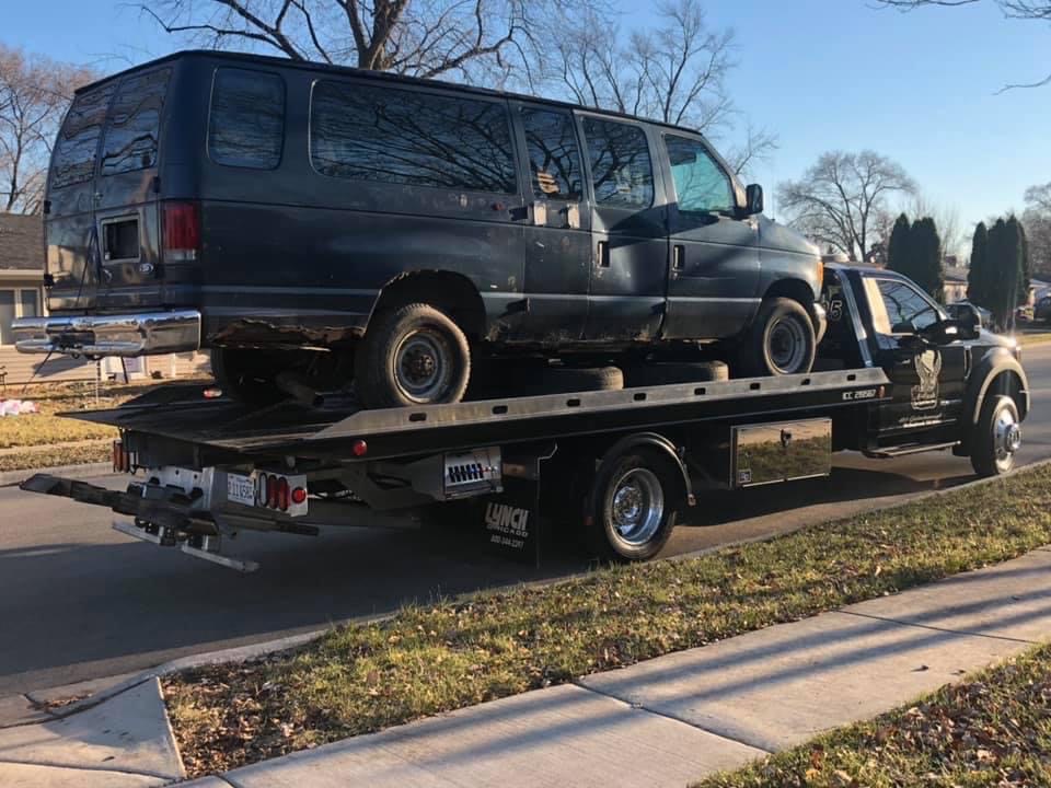 A tow truck on the street towing away a black junk van