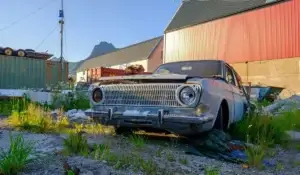 Old junk car parked in a scrapyard with mountains and industrial buildings in the background