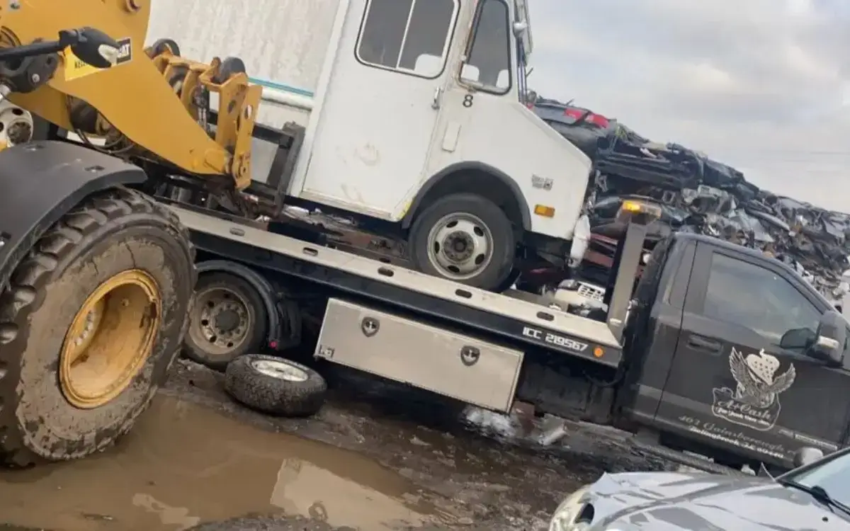 Loader lifting a junk truck onto a flatbed surrounded by stacked junk cars in a salvage yard.